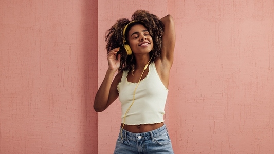 Beautiful woman with curly hair enjoying music with closed eyes against pink wall