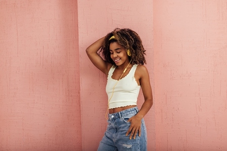 Cheerful woman with curly hair leaning pink wall while listening music