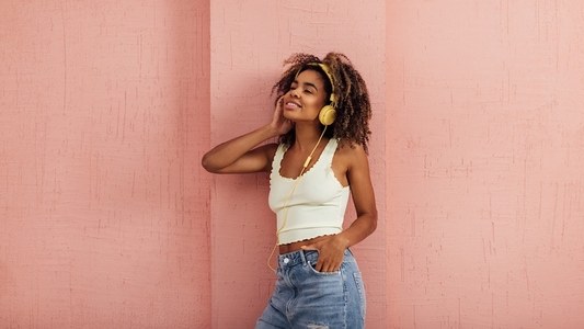 Young female in casuals leaning on a pink wall listening to music by yellow headphones