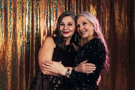 Two smiling senior girlfriends wearing evening dresses hugging each other while standing at glitter background