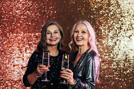 Stylish senior females standing together against glitter backdrop holding glasses of champagne