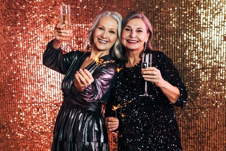 Two happy senior women holding sparklers and glasses of champagne against a glitter background