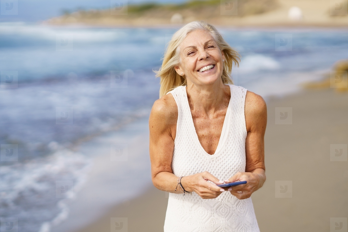 Smiling senior female walking on the beach using a smartphone