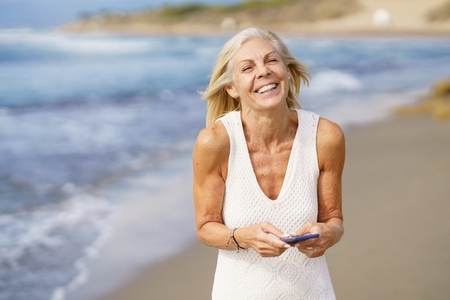 Smiling senior female walking on the beach using a smartphone