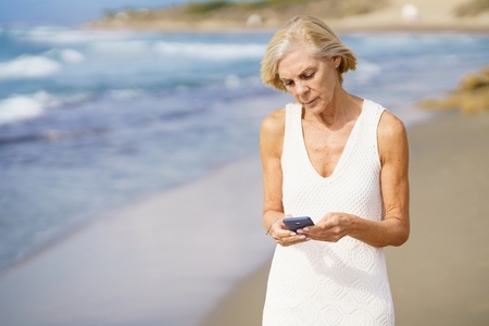 Senior woman walking on the beach using a smartphone