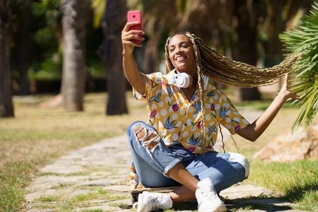 Black woman with braided hair taking a selfie with a smartphone sitting on her skateboard