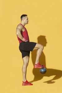 Athletic man in sportswear lifting kettle bell with foot against yellow background