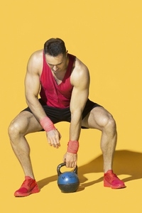 Strong athletic man squatting and lifting kettle bell against yellow background