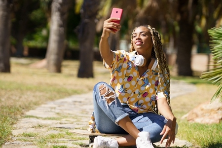 African female with braided hair taking a selfie with a smartphone sitting on her skateboard