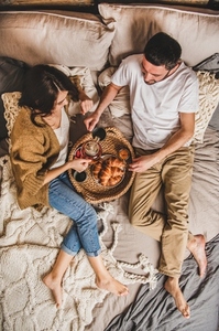 Young happy couple lying in bed barefoot and having breakfast
