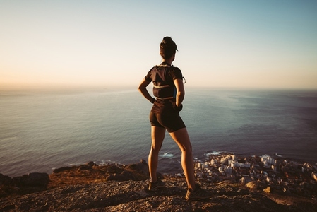 Back view of a woman in hiking sportswear standing on a cliff at sunset  Sportswoman looking at the ocean on the top of the mountain