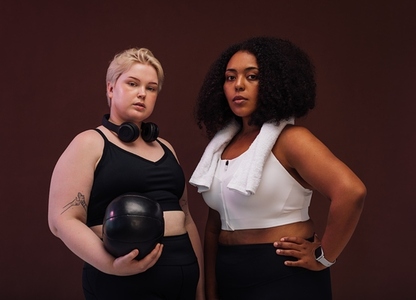 Two plus size women in sports clothes standing in studio  Young female athletes posing together against brown background