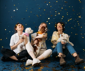 Three laughing women with their dogs sitting on a blue background under colorful confetti
