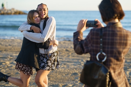 Faceless woman taking photo of hugging diverse girlfriends on seashore