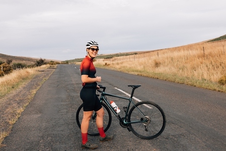 Smiling sportswoman wearing a cycling helmet and sunglasses standing with a bicycle on an empty countryside road