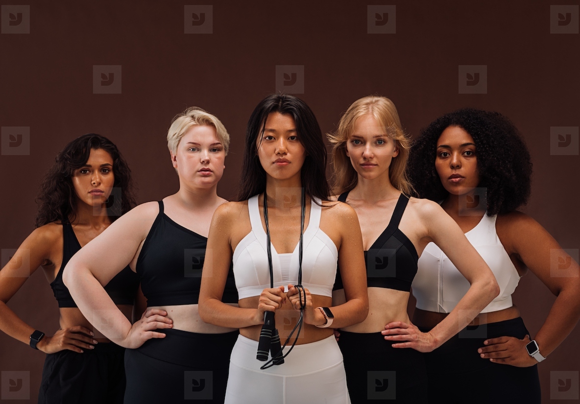 Five women in sportswear looking at camera  Females with different body types and ethnicities standing together against brown background