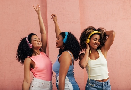 Three girlfriends with different body types dancing together  Smiling females with headphones enjoying music at pink wall