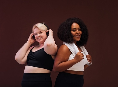 Two smiling plus size women standing together in studio  Females in sportswear posing together against brown background