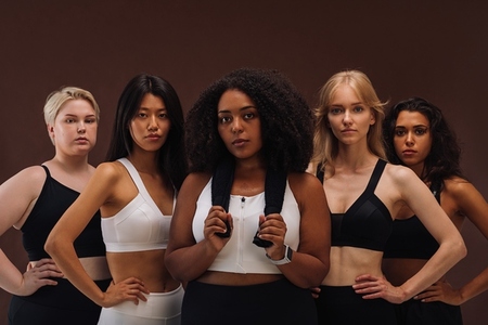 Diverse women in sportswear looking at the camera while standing against a brown background  Five females of different races in studio
