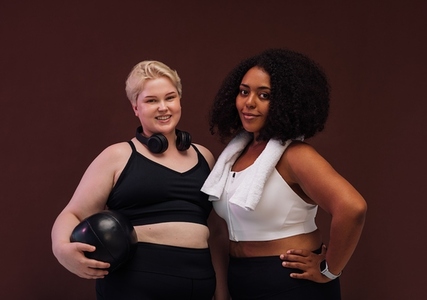 Two cheerful women in sportswear posing on brown background  Plus size females standing together with fitness equipment
