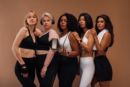 Five diverse females in sports clothes with fitness accessories standing together in studio