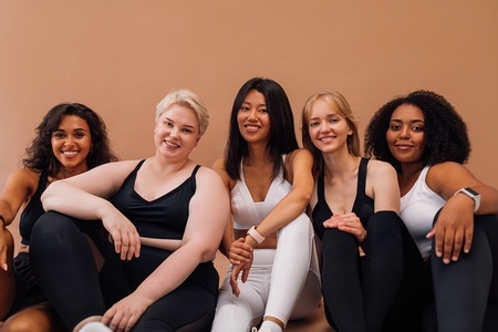 Five smiling women in fitness clothes sitting together on pastel background  Females with different body types relaxing after workout
