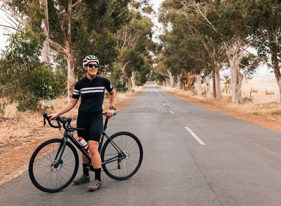 Female cyclist in sportswear wearing sunglasses and helmet relaxing after ride standing on empty road