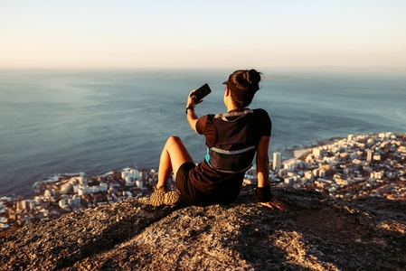 Back view of sportswoman relaxing during trail run  Fit woman sitting on the top of the mountain and taking selfie at sunset
