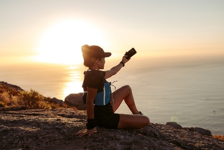 Young fit woman in sportswear taking selfie while sitting on a hill at sunset
