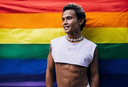 Smiling young gay man in stylish clothes standing against pride flag