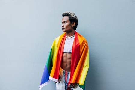 Handsome young man with pride movement LGBT flag on shoulders against grey wall outdoors