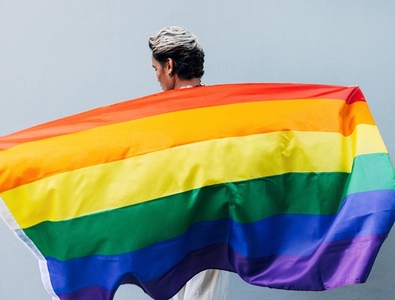 Back view of a young man holding a LGBT rainbow flag at grey wall outdoors