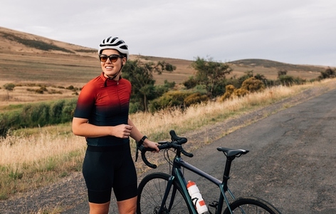 Smiling woman cyclist wearing sunglasses and helmet taking a break during ride standing on a road