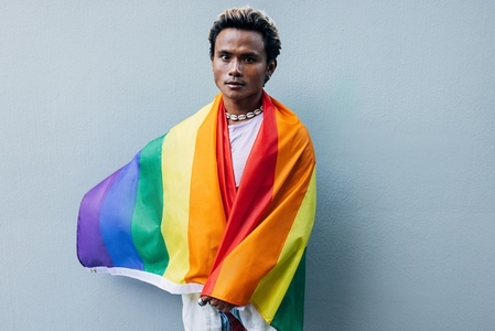 Young man wrapped in rainbow LGBT flag standing at grey wall outdoors