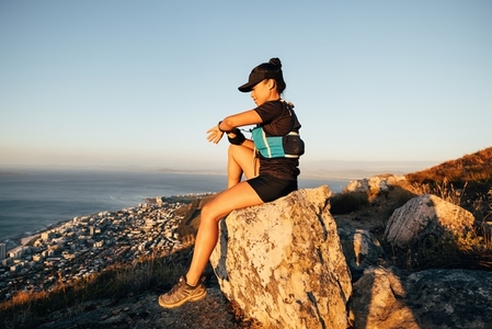 Side view of sports woman sitting on a rock checking smart watch at sunset  Trail runner relaxing on the top of the mountain