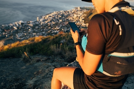 Close up woman hiker in sportsclothes checking smartphone while sitting on the top of the mountain at sunset