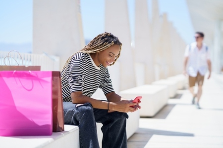 Black woman using her smartphone sitting on a bench in the street
