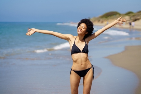 Positive African American female opening her arms on the beach to enjoy her holiday in the sun