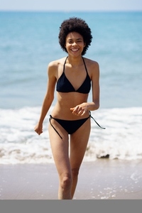 Happy black woman laughing while walking in bikini on the sand of the beach