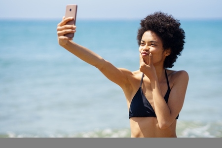 Black woman with afro hairstyle taking a selfie with a funny gesture on a tropical beach