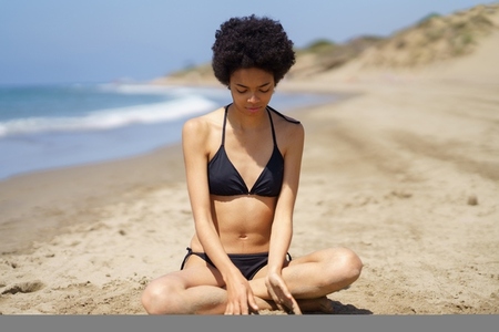 Positive black girl in bikini sitting on the sand of a beach gazing thoughtfully at the sand