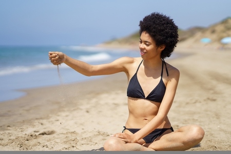 Positive black woman in bikini sitting playing with sand on the beach