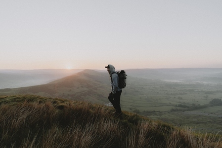 Male hiker with backpack on hill overlooking rural landscape at sunrise