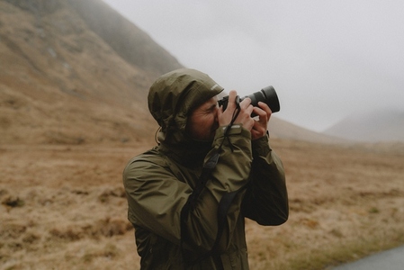 Male photographer in hooded jacket using camera below mountain