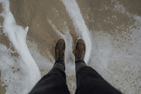 POV legs of man in jeans and boots standing in ocean sea foam on sandy beach