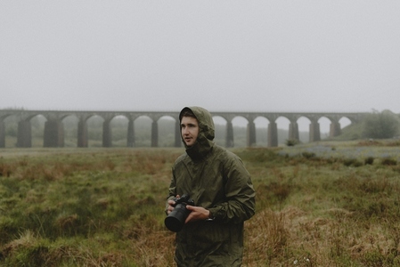 Male photographer in jacket with camera in rainy field with aqueduct