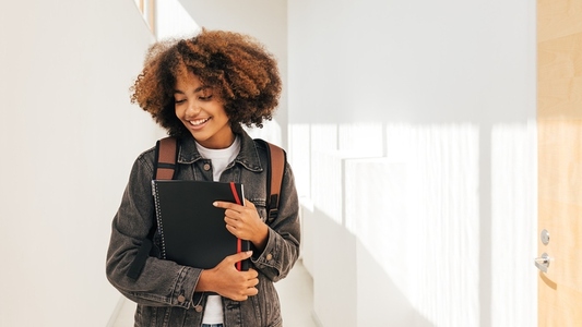 Cheerful student with curly hair standing in hallway  Girl holding notebook and looking down