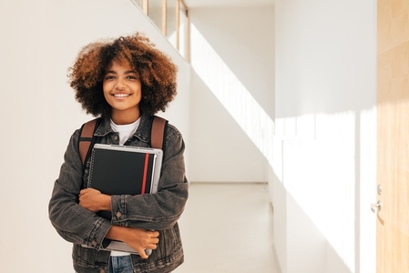 Portrait of a smiling student holding notebook and laptop while standing in corridor