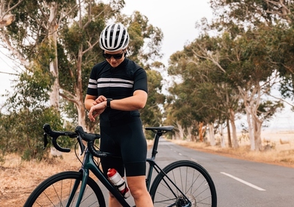 Professional woman cyclist standing with her bike on empty road checking fitness tracker