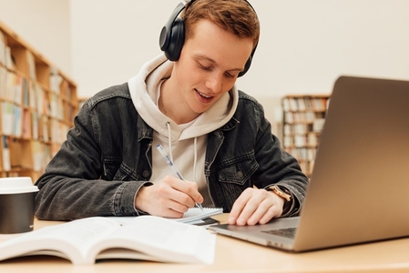 Male student preparing exams at desk in library  Smiling guy in headphones writing on notebook